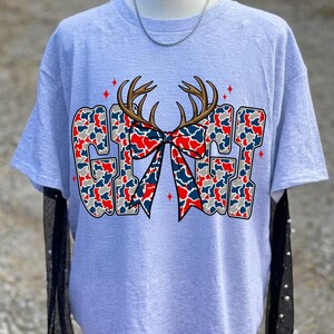 May include: A light gray short-sleeve t-shirt featuring the text GIGI in a red, white, and blue camouflage pattern. The design includes a brown deer antler graphic centered above a matching camouflage bow, accented by small red four-pointed stars.