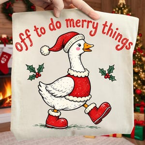 May include: A cream-coloured t-shirt featuring a graphic of a white goose wearing a red Santa hat, a red vest with white trim, and red boots. The text "off to do merry things" is printed in red above the goose, flanked by two holly sprigs.