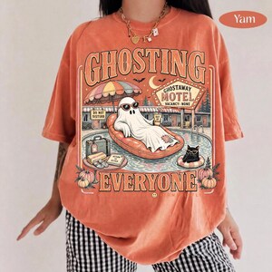 Puede incluir: Una camiseta naranja con un estampado gr&aacute;fico que incluye el texto GHOSTING EVERYONE. El dise&ntilde;o muestra un fantasma con gafas de sol descansando en un flotador de piscina en un motel, junto a un gato negro, calabazas y un letrero.