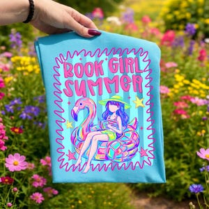 May include: A light blue t-shirt featuring a graphic print with the text BOOK GIRL SUMMER in pink. The design includes a person wearing a hat and reading a book while sitting on a large, colourful flamingo pool float, surrounded by stars.