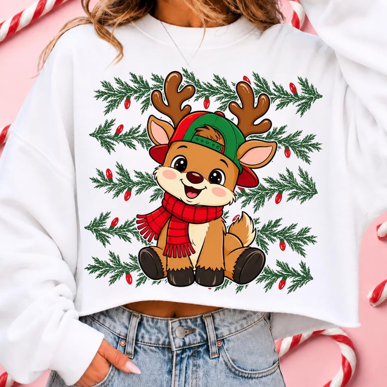 May include: A white cropped sweatshirt featuring a graphic of a cartoon reindeer wearing a green baseball cap and a red scarf. The background of the graphic consists of horizontal rows of green pine branches interspersed with small red light bulbs.