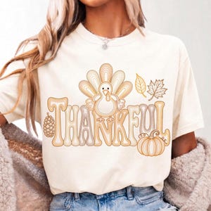 May include: A cream-colored t-shirt featuring a brown line-art graphic of a turkey, a pinecone, a pumpkin, and autumn leaves. The word THANKFUL is printed in large, stylized, multi-layered brown lettering integrated with the turkey illustration.
