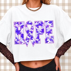 May include: A white short-sleeved t-shirt featuring the word HOPE in large block letters, with the letter O replaced by a purple awareness ribbon. The letters have a purple and light blue camouflage pattern with gold speckles throughout.