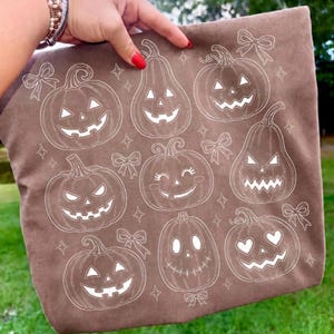 May include: A brown tote bag featuring a white line art pattern of various jack-o'-lanterns with different facial expressions, including smiling, heart-shaped eyes, and jagged teeth, interspersed with small decorative bows and four-pointed stars.