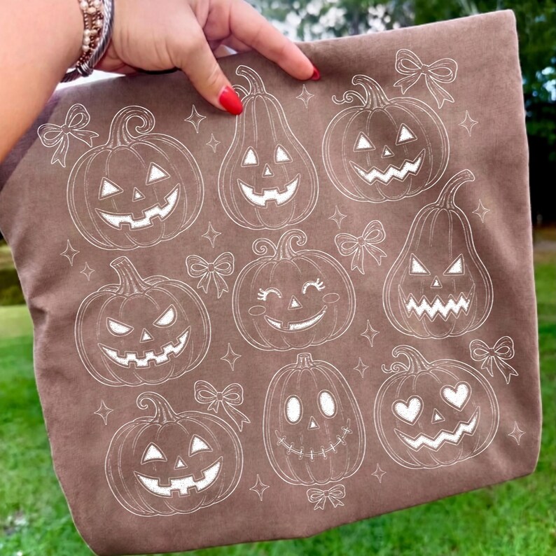 May include: A brown tote bag featuring a white line art pattern of various jack-o'-lanterns with different facial expressions, including smiling, heart-shaped eyes, and jagged teeth, interspersed with small decorative bows and four-pointed stars.