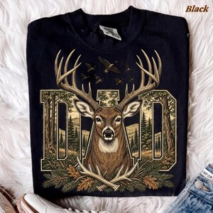 May include: A black short-sleeve t-shirt featuring a large graphic print of a deer head with antlers centred within the block letters DAD. The design includes a forest landscape background with pine trees and flying birds, plus crossed antlers.