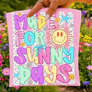 May include: A light pink fabric pouch featuring a graphic print with the text MADE FOR SUNNY DAYS in colorful, patterned bubble letters. The design includes a yellow smiley face sun, small blue starfish, and various flowers over vertical stripes.