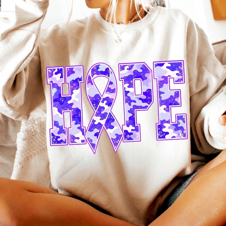 May include: A beige crewneck sweatshirt featuring the word HOPE printed in large block letters. The letter O is replaced by an awareness ribbon. The text has a purple and light blue camouflage pattern with a distressed, textured finish.