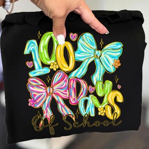 Op de afbeelding: Zwarte hoodie met een kleurrijke afbeelding ter viering van 100 schooldagen. Het ontwerp toont het nummer "100", strikken, harten en de woorden "Days of School" in een speels lettertype. Het ontwerp is in heldere kleuren.
