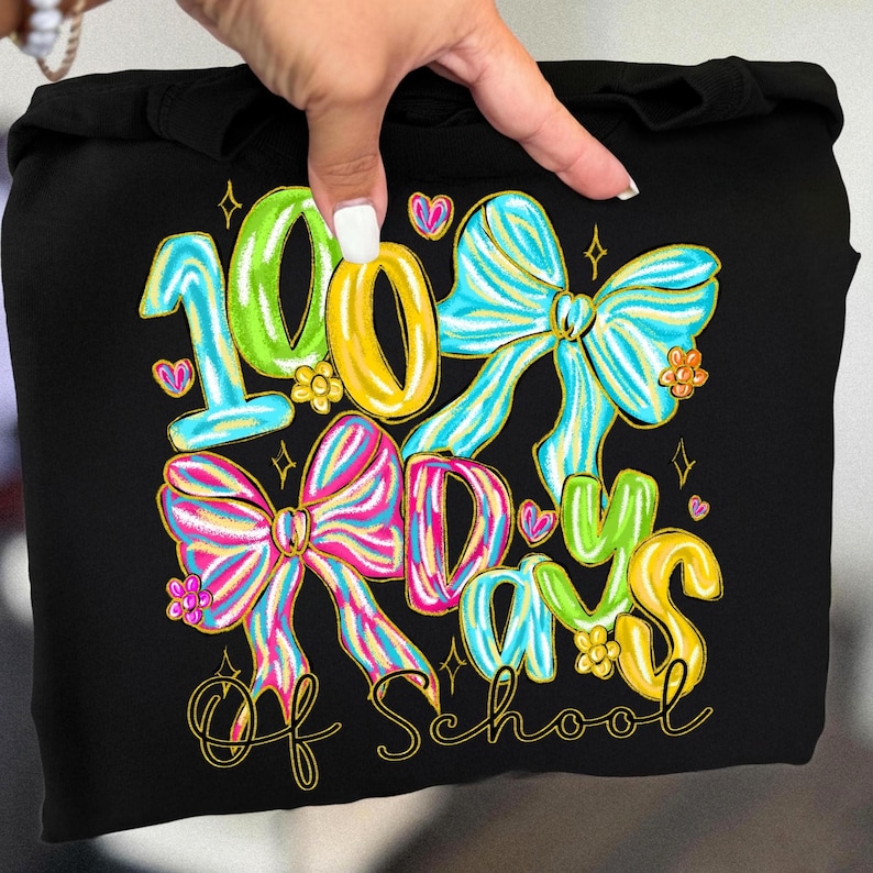 Op de afbeelding: Zwarte hoodie met een kleurrijke afbeelding ter viering van 100 schooldagen. Het ontwerp toont het nummer "100", strikken, harten en de woorden "Days of School" in een speels lettertype. Het ontwerp is in heldere kleuren.