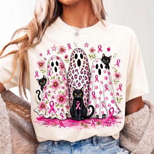 May include: A cream-colored t-shirt featuring a graphic of three ghosts and three black cats. The ghosts are decorated with pink flowers, leopard print, and pink polka dots, all accented with pink awareness ribbons, stars, and a pink base.