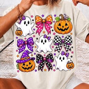 May include: A cream-coloured t-shirt featuring a repeating pattern of white ghosts, orange jack-o'-lanterns, and various decorative bows in polka dot, checkered, and solid patterns, accented with small purple and orange stars throughout the design.