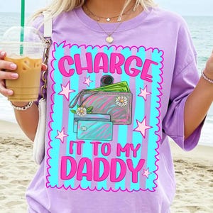 May include: A light purple t-shirt featuring a graphic with a blue and purple striped background, pink stars, and a wallet with cash and credit cards. The text reads "CHARGE IT TO MY DADDY" in bold pink letters with a scalloped pink border.