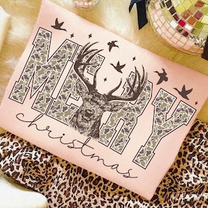 May include: A light pink t-shirt with the words "MERRY Christmas" in a camouflage pattern. A detailed illustration of a deer is centered on the shirt, with several flying birds and stars. The shirt is on a leopard print surface.