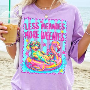 May include: A light purple t-shirt featuring a graphic of a dachshund wearing heart-shaped sunglasses while floating on a pink flamingo pool float. The text reads "LESS MEANIES MORE WEENIES" in bright pink letters against a blue and purple striped background.