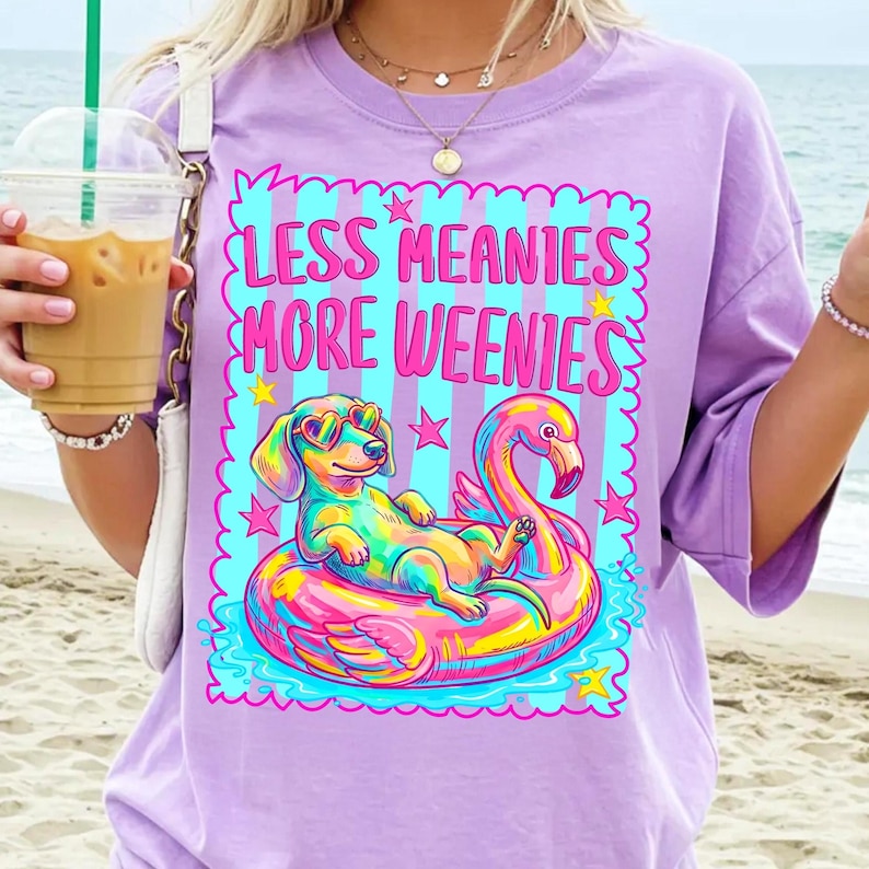 May include: A light purple t-shirt featuring a graphic of a dachshund wearing heart-shaped sunglasses while floating on a pink flamingo pool float. The text reads "LESS MEANIES MORE WEENIES" in bright pink letters against a blue and purple striped background.