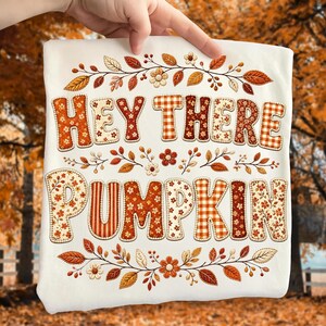 May include: A white t-shirt featuring the text HEY THERE PUMPKIN in large, block-style letters filled with various orange, brown, and cream autumn-themed patterns, surrounded by decorative illustrations of fall leaves and small flowers.