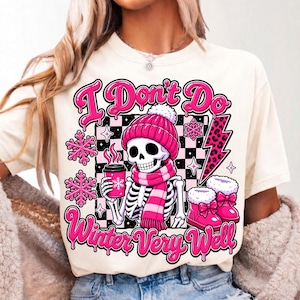 May include: A cream-coloured t-shirt featuring a graphic of a skeleton wearing a pink beanie and scarf, holding a pink coffee cup. The design includes pink snowflakes, a leopard print lightning bolt, and the text "I Don't Do Winter Very Well."