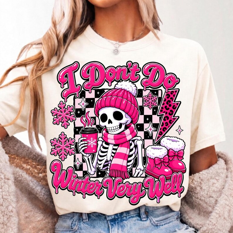 May include: A cream-coloured t-shirt featuring a graphic of a skeleton wearing a pink beanie and scarf, holding a pink coffee cup. The design includes pink snowflakes, a leopard print lightning bolt, and the text "I Don't Do Winter Very Well."