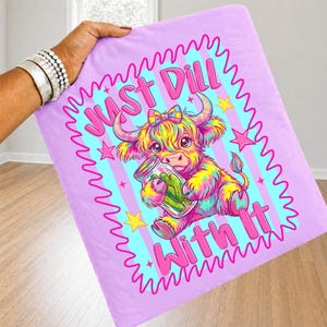 Puede incluir: Un cuadrado de tela de color morado claro con una ilustraci&oacute;n colorida de una vaca highland con un lazo, sosteniendo un frasco de cristal con pepinillos. El texto JUST DILL WITH IT aparece en letras rosas, rodeado por un borde rosa ondulado y estrellas amarillas.
