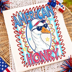 May include: A white t-shirt featuring a graphic of a goose wearing a baseball cap and sunglasses. The text reads AMERICAN HONRY in blue and pink letters. The design includes a red wavy border, blue and yellow stars, and light blue stripes.