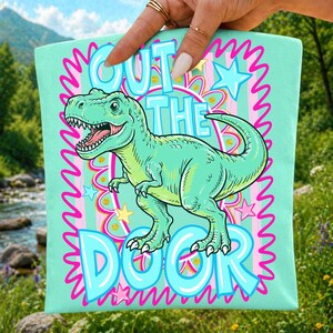 May include: A light mint green fabric square featuring a central illustration of a green Tyrannosaurus Rex. The text reads OUT THE DOOR in large blue letters. The design includes pink squiggly borders, yellow and pink stars, and circles.