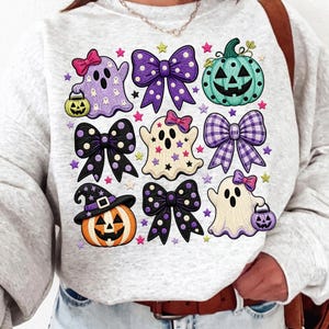 K&ouml;nnte beinhalten: Ein hellgrau meliertes Sweatshirt mit einem Grafikdruck aus drei Geistern, drei K&uuml;rbissen und vier Schleifen. Die Geister sind wei&szlig; und lila, die K&uuml;rbisse orange und blaugr&uuml;n, und die Schleifen sind lila, schwarz und kariert mit Sternakzenten.