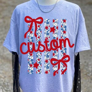 May include: A light gray t-shirt featuring a graphic design of four vertical stripes with a red and blue floral pattern. The design includes two red bows, two red stars, and the word custom written in red cursive script across the center.