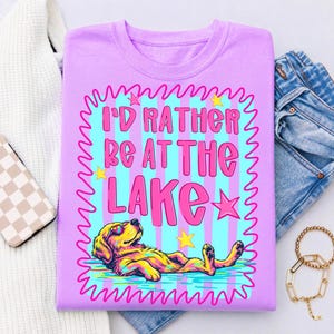 May include: A light purple t-shirt featuring a graphic with the text I'D RATHER BE AT THE LAKE in pink letters. The design includes a colorful illustration of a dog wearing sunglasses lying down, surrounded by pink stars and a wavy border.