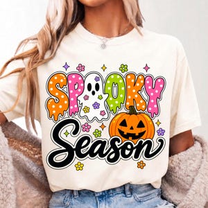 May include: A cream-colored t-shirt featuring a colorful graphic with the text "SPOOKY SEASON". The design includes a white ghost, an orange jack-o'-lantern, and various small flowers and stars in pink, orange, green, and purple hues.