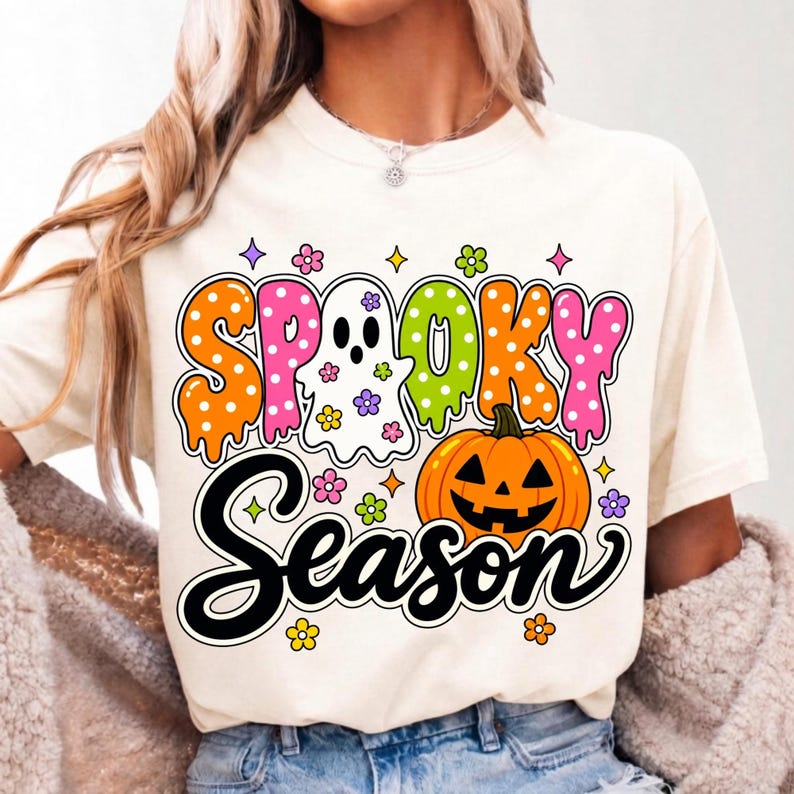 May include: A cream-colored t-shirt featuring a colorful graphic with the text "SPOOKY SEASON". The design includes a white ghost, an orange jack-o'-lantern, and various small flowers and stars in pink, orange, green, and purple hues.