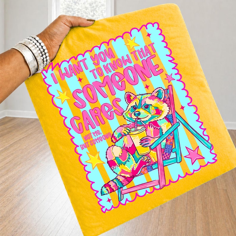 K&ouml;nnte beinhalten: Ein gelbes Dekokissen mit einer bunten Grafik eines Waschb&auml;ren, der in einem Liegestuhl sitzt und aus einem Becher trinkt. Der Text lautet: I WANT YOU TO KNOW THAT SOMEONE CARES NOT ME, BUT SOMEONE. Das Design enth&auml;lt Sterne und einen gewellten Rand.