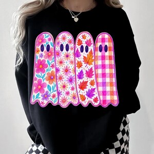 May include: A black long-sleeve sweatshirt featuring a graphic of four ghosts standing side-by-side. Each ghost has a unique pattern: one with large pink and orange flowers, one with small daisies, one with autumn leaves, and one with a pink and orange plaid.