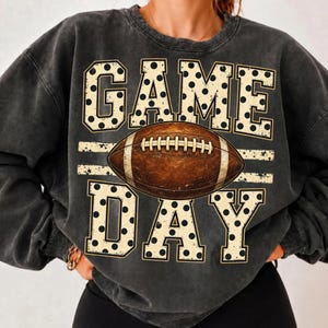 Op de afbeelding: Een antracietgrijze sweater met ronde hals en een grafische print met de tekst GAME DAY in grote, cr&egrave;mekleurige blokletters met zwarte stippen. Een bruine American football staat centraal tussen de woorden GAME en DAY.
