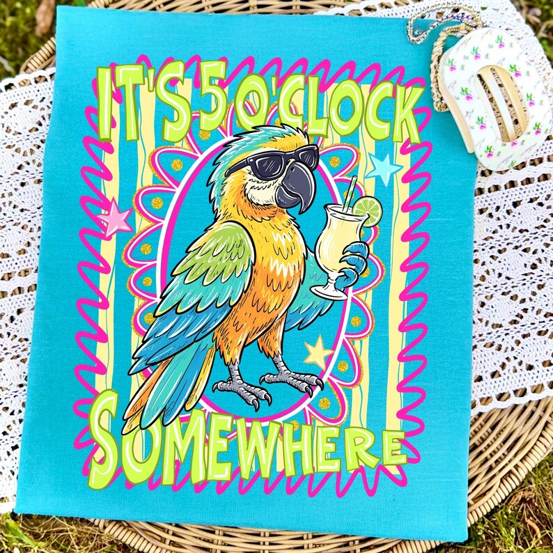 May include: A turquoise towel featuring a cartoon parrot wearing sunglasses and holding a cocktail. The text "IT'S 5 O'CLOCK SOMEWHERE" is in neon green and pink. The parrot is yellow, blue, and orange.