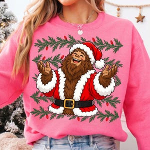 Puede incluir: Una sudadera de cuello redondo en color rosa brillante con un gr&aacute;fico de un Bigfoot marr&oacute;n vestido con un traje de Pap&aacute; Noel rojo con ribete de piel blanca y un cintur&oacute;n negro. El dise&ntilde;o est&aacute; rodeado de ramas de pino verdes y bombillas rojas.