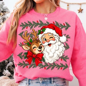 May include: A bright pink crewneck sweatshirt featuring a graphic print of a winking Santa Claus and a smiling reindeer with a large red bow. The design is framed by green pine branches and small red lights, creating a festive holiday aesthetic.