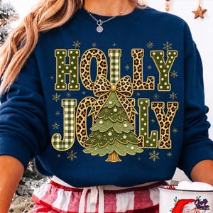 May include: A navy blue crewneck sweatshirt featuring the text HOLLY JOLLY in a mix of green polka dot, green plaid, and leopard print patterns. A central green Christmas tree graphic with a leopard print bow sits between the words.