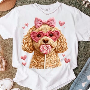 May include: White t-shirt featuring a detailed illustration of a poodle wearing heart-shaped sunglasses and a pink bow. The poodle is holding a lollipop. Pink hearts are scattered around the design. The shirt has a crew neck and short sleeves.
