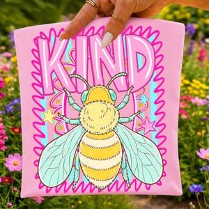Pode incluir: Um artigo de tecido rosa claro com uma grande ilustra&ccedil;&atilde;o de uma abelha amarela e azul clara sob a palavra KIND em letras rosa em negrito. O design inclui uma borda rosa decorativa, listras verticais azul claras e pequenas estrelas.