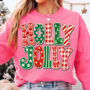May include: A bright pink crewneck sweatshirt featuring the text HOLLY JOLLY in large, stylized letters. Each letter displays a different festive pattern, including red bows, green polka dots, red gingham, green Christmas trees, and red candy stripes.