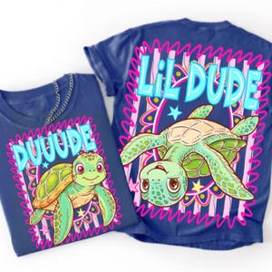 May include: A dark blue t-shirt featuring a graphic of a green sea turtle. The smaller design on the left says DUUUDE in light blue letters, while the larger design on the right says LIL DUDE in light blue letters, both framed by pink borders.