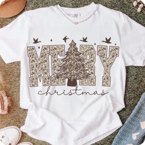 May include: White t-shirt with the words "MERRY CHRISTMAS" in a camouflage pattern, a brown Christmas tree, and flying birds. The word "christmas" is written in a cursive font below the main text.