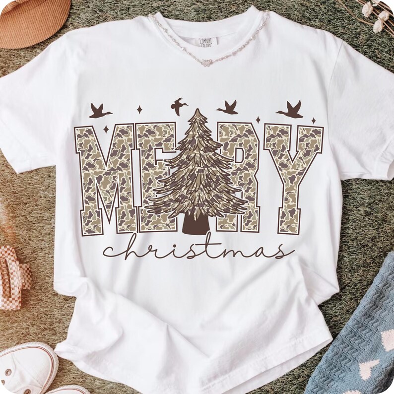 May include: White t-shirt with the words "MERRY CHRISTMAS" in a camouflage pattern, a brown Christmas tree, and flying birds. The word "christmas" is written in a cursive font below the main text.