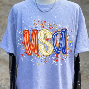 Może przedstawiać: Jasnoszary t-shirt z kr&oacute;tkim rękawem z napisem USA dużymi, stylizowanymi literami. Litera U jest czerwona, S biała, a A niebieska. Wz&oacute;r otoczony jest żywym motywem rozprysk&oacute;w czerwonych, białych, niebieskich i złotych kropek farby.