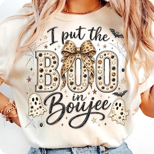 Pu&ograve; includere: Una t-shirt color crema con una stampa grafica che riporta il testo I put the BOO in Boujee. Il design include un grande fiocco con stampa leopardata, due fantasmi, ragni, ragnatele, pipistrelli e stelle nei toni del nero, oro e marrone.
