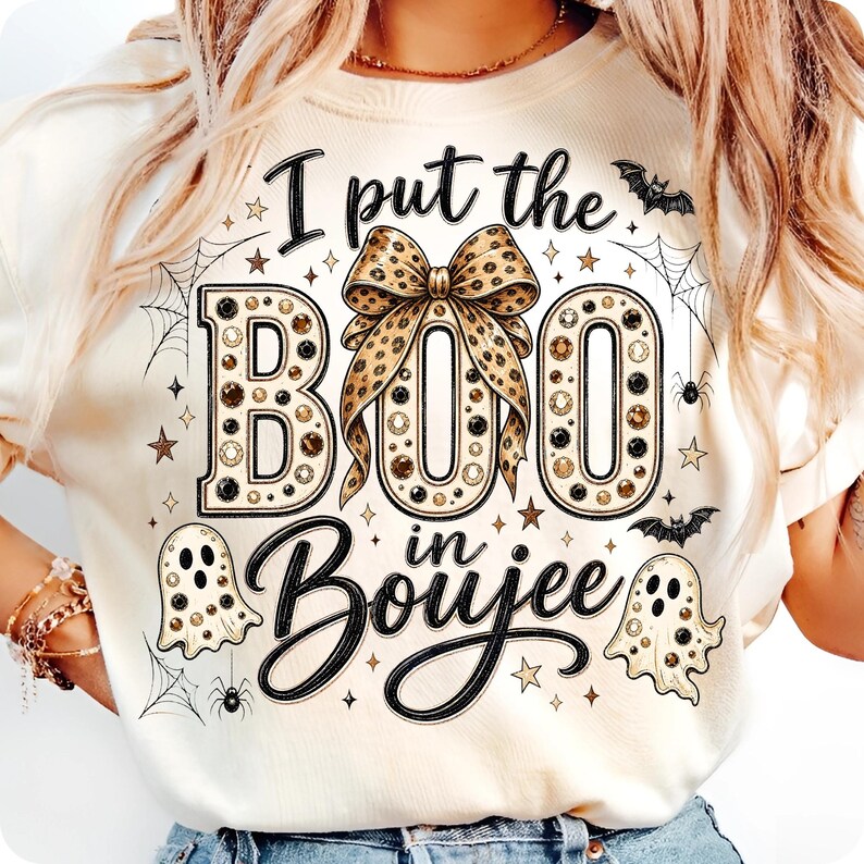 Pu&ograve; includere: Una t-shirt color crema con una stampa grafica che riporta il testo I put the BOO in Boujee. Il design include un grande fiocco con stampa leopardata, due fantasmi, ragni, ragnatele, pipistrelli e stelle nei toni del nero, oro e marrone.