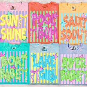 May include: Six colorful t-shirts with summer-themed text designs. The shirts are pink, orange, yellow, teal, light blue, and lavender. Each shirt features a different phrase in bold, colorful letters, such as "Sun Shine" and "Pool Bum".