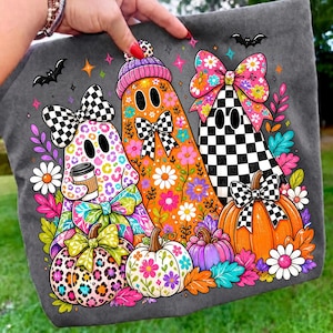 Puede incluir: Una camiseta gris oscuro con un gr&aacute;fico de tres fantasmas decorados con patrones coloridos, incluyendo estampado de leopardo, dise&ntilde;os florales naranjas y cuadros blancos y negros, rodeados de calabazas, flores y murci&eacute;lagos.