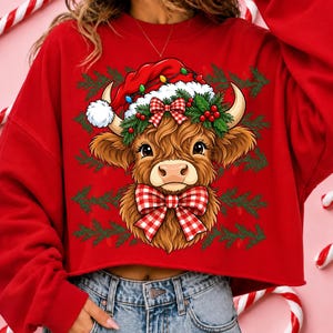 Puede incluir: Una sudadera corta de color rojo brillante con el gr&aacute;fico de una vaca Highland marr&oacute;n y peluda que lleva un gorro de Pap&aacute; Noel rojo con luces navide&ntilde;as, una corona de acebo verde y un gran lazo a cuadros rojos y blancos sobre ramas de pino.